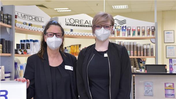Die beiden Frauen tragen einen Mund-Nasen-Schutz und stehen vor mehreren Friseurutensilien, die in einem Regal liegen.