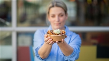 Eine Frau im blauen Hemd hält eine Pappschale mit Pommes und Avocadocreme ins Bild. 