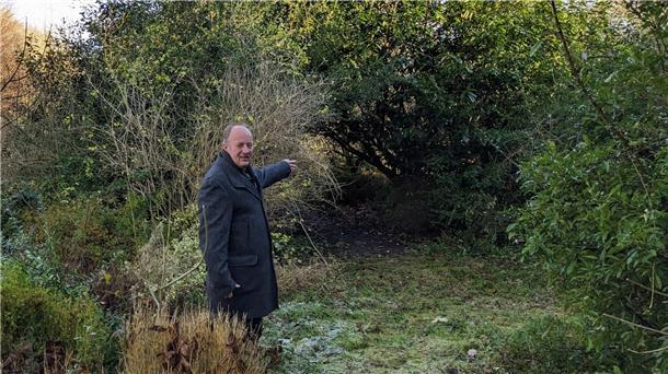 Ein Mann im dunklen Mantel steht in einem überwucherten Gartenbereich und zeigt auf ein zugewachsenes, dichtes Gebüsch.