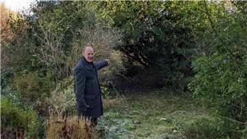 Ein Mann im dunklen Mantel steht in einem überwucherten Gartenbereich und zeigt auf ein zugewachsenes, dichtes Gebüsch.
