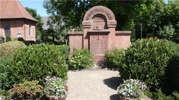 Ein Kriegerdenkmal im HintergrundDrumherum Sträucher und Blumenkübel.