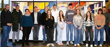 Eine Gruppe aus Jugendlichen und Erwachsenen steht vor einem Schul-Kiosk mit bunten Fenstern. Einige tragen lässige Kleidung, andere formelle Outfits. Im Hintergrund hängen Schilder zur Neueröffnung des Kiosks.