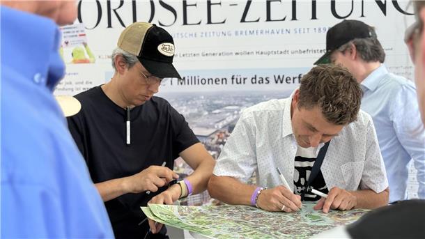 Zwei Männer stehen vor einem Poster, das auf einem hohen Tisch liegt. Einer von ihnen zeichnet darauf.