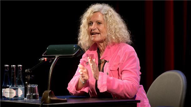 Eine Frau in rosa Bluse sitzt auf der Bühne und liest vor
