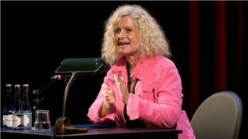 Eine Frau in rosa Bluse sitzt auf der Bühne und liest vor