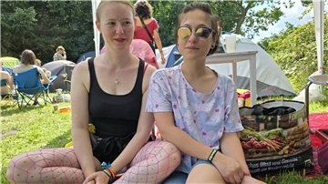 Zwei Frauen sitzen im Gras, im Hintergrund Menschen in Campingstühlen
