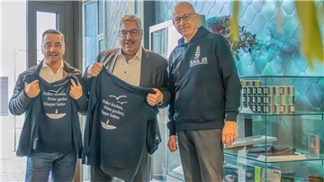 Drei Männer stehen nebeneinander in einem modernen Raum mit blauem Wabenmuster an der Wand. Zwei Männer halten schwarze Pullover mit der Aufschrift "Poller hocken, Pötte gucken, Klappe halten" und einer Grafik eines Bootes auf der Rückseite. Der dritte Mann trägt ebenfalls einen dunkelblauen Pullover mit der Aufschrift "Sail 25 Bremerhaven" auf der Vorderseite.