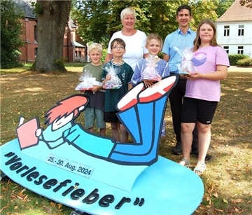 Zwei Erwachsene und vier Kinder mit Geschenken stehen auf einer Grünfläche vor einem Aufsteller, auf dem "Vorlesefieber" und "25.-30. Aug. 2024" steht.