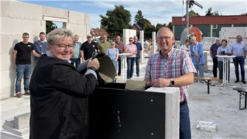 Eine Frau kippt einen Eimer mit Beton in eine Vorrichtung. Gegenüber steht ein lächelnder Mann in Hemd mit einer Kelle voller Beton.