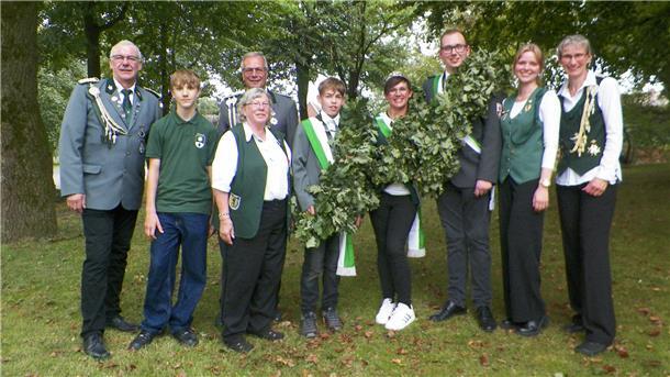 Neun Schützinnen und Schützen in Uniform stehen nebeneinander auf einer Wiese zwischen Bäumen.
