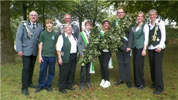 Neun Schützinnen und Schützen in Uniform stehen nebeneinander auf einer Wiese zwischen Bäumen.