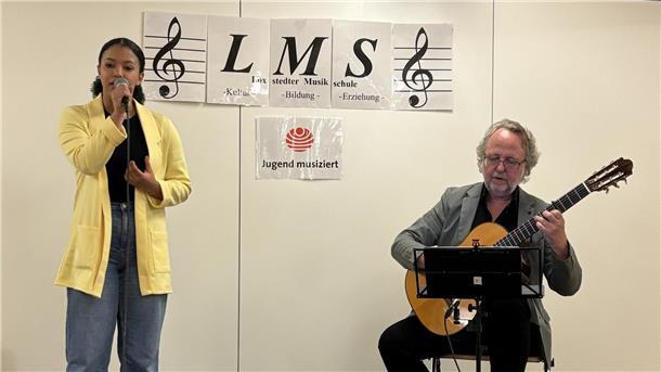 Loxstedter Musikschüler feiern ihre großen Erfolge bei „Jugend musiziert“ 2025 Eine junges Mädchen singt in ein Mikrofon, während ein älterer Mann sie auf einer klassischen Gitarre begleitet. Im Hintergrund hängen Schilder mit der Aufschrift „LMS – Loxstedter Musikschule“ sowie das Logo von „Jugend musiziert“.