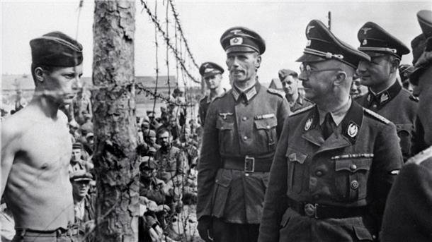SS-Chef Heinrich Himmler blickt einen Kriegsgefangenen hinter einem Zaun an.