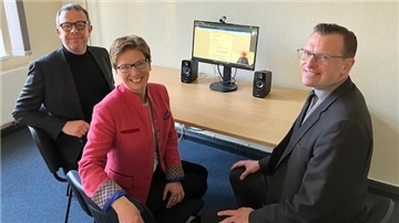 Zwei Herren (links und rechts) lächeln, eine Frau (Mitte) auch. Auf einem Schreibtisch ist ein Bildschirm zu sehen, der eingeschaltet ist. Der Start der Online-Rechtsantragstelle im Arbeitsgericht Bremen-Bremerhaven erleichtert den Bürgern den Zugang zur Justiz, indem sie Anträge und Klagen bequem von zuhause aus einreichen können.