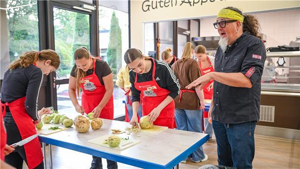 Jugendliche schneiden Gemüse, während ein Mann Anweisungen gibt.