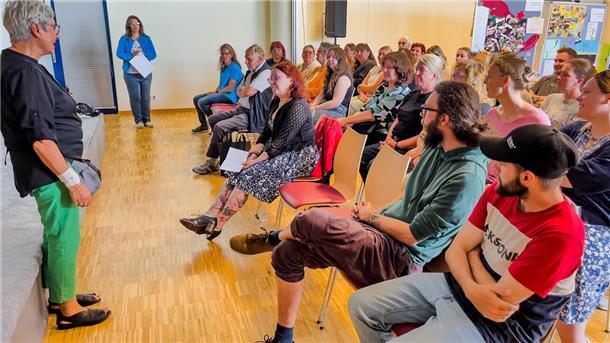 Kita Am Oberhamm gewinnt Wettbewerb mit Regenwurm-Projekt Eine Gruppe von Erwachsenen sitzt in einem Veranstaltungsraum und hört einer Frau im Vordergrund aufmerksam zu. Die Frau steht seitlich zur Kamera und spricht zu den Teilnehmenden. Im Hintergrund sind weitere Personen sowie Plakate und Infowände zu sehen.