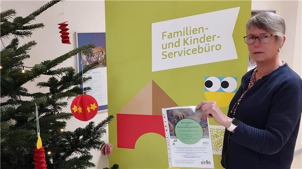 Links ist ein Weihnachtsbaum zu sehen, rechts eine Frau, die einen Zettel in die Kamera hält. Im Hintergrund steht ein Aufsteller.