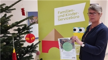 Links ist ein Weihnachtsbaum zu sehen, rechts eine Frau, die einen Zettel in die Kamera hält. Im Hintergrund steht ein Aufsteller.