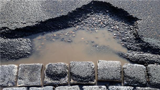 Ein mit Wasser gefülltes Schlagloch in einer Asphaltstraße neben gepflasterten Steinen.