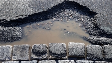 Ein mit Wasser gefülltes Schlagloch in einer Asphaltstraße neben gepflasterten Steinen.