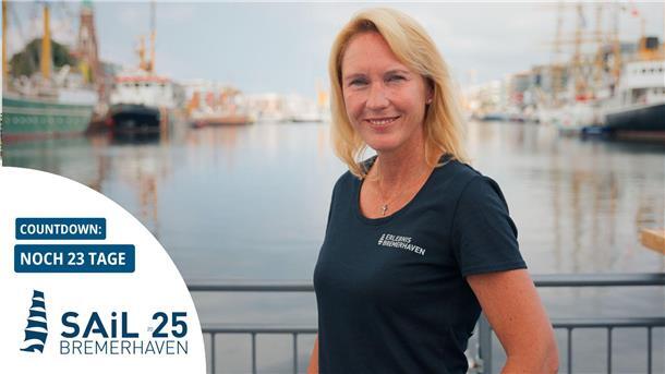 So hält Tanja Mehl die besonderen Momente der Sail 2025 fest Frau im T-Shirt mit „Erlebnis Bremerhaven“-Logo steht lächelnd am Hafen; im Hintergrund liegen historische Segelschiffe. Links im Bild Countdown zur Sail 2025 Bremerhaven: Noch 23 Tage.