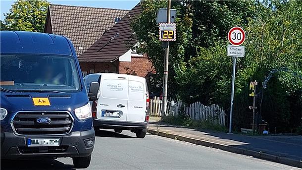 Warum in Langen auf der Landesstraße 120 immer noch Tempo 30 gilt Zwei Lieferwagen fahren auf einer Straße mit Tempo-30-Zone in einem Wohngebiet. Rechts steht ein Verkehrsschild mit der Aufschrift „30 – Straßenschäden“ sowie eine digitale Geschwindigkeitsanzeige, die „22“ km/h zeigt.