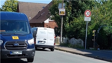 Zwei Lieferwagen fahren auf einer Straße mit Tempo-30-Zone in einem Wohngebiet. Rechts steht ein Verkehrsschild mit der Aufschrift „30 – Straßenschäden“ sowie eine digitale Geschwindigkeitsanzeige, die „22“ km/h zeigt.