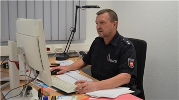Ein Mann in Polizeiuniform mit kurzen dunklen Haaren sitzt vor einem Computer und schaut auf den Bildschirm. 