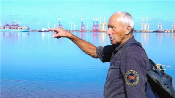 Ein älterer Mann mit Rucksack zeigt über das Wasser auf Containerterminals und Windräder am gegenüberliegenden Ufer; im Hintergrund ist ein Hafen mit Kränen sichtbar.