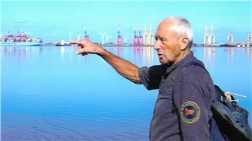Ein älterer Mann mit Rucksack zeigt über das Wasser auf Containerterminals und Windräder am gegenüberliegenden Ufer; im Hintergrund ist ein Hafen mit Kränen sichtbar.