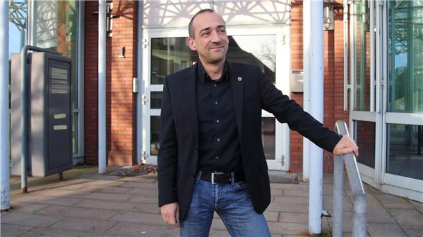 Thomas Lötsch steht vor dem Eingang eines Gebäudes. Er hält eine Hand an einem kurzen Treppengeländer. Er trägt Jeans, dunkles Hemd und dunkles Sakko.