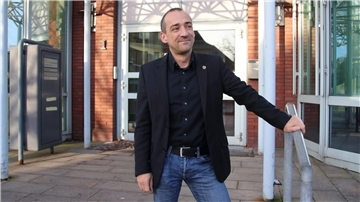 Thomas Lötsch steht vor dem Eingang eines Gebäudes. Er hält eine Hand an einem kurzen Treppengeländer. Er trägt Jeans, dunkles Hemd und dunkles Sakko.