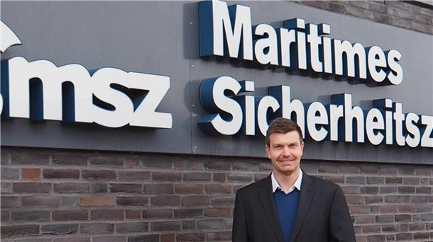 Ein Mann in einem dunklen Anzug und blauen Pullover steht lächelnd vor einer Backsteinwand mit einem Schild, auf dem „Maritimes Sicherheitszentrum“ steht.