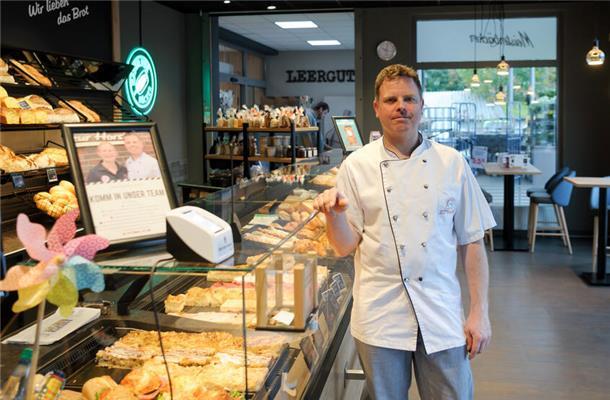 Ein Mann mit weißem Kittel vor einer Theke mit Kuchen, Brot und Brötchen. Er steht in einer Bäckerfiliale.