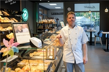Ein Mann mit weißem Kittel vor einer Theke mit Kuchen, Brot und Brötchen. Er steht in einer Bäckerfiliale.
