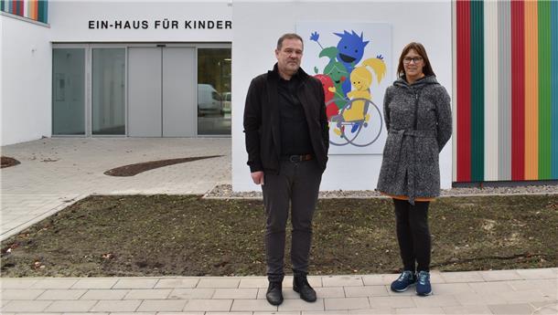 Zwei Personen stehen vor einer Wand mit einem großen bunten Logo und bunten Streifen.