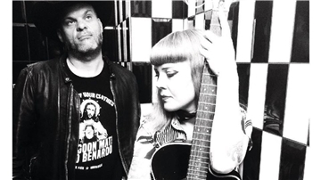 Ein Mann steht im Hintergrund. Im Vordergrund hält eine Frau eine Gitarre im Arm.