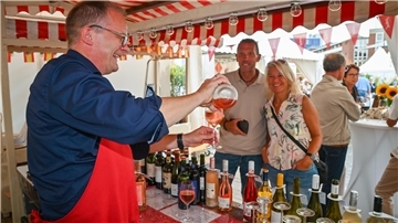 Ein Mann schenkt in einem Verkaufsstand Wein in ein Glas. Im Hintergrund stehen ein weiterer Mann und eine Frau und warten darauf, den Wein in Empfang zu nehmen.