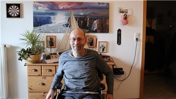 Mann mit Brille im Rollstuhl vor Wohnzimmer-Wand