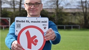 TSV-Vorsitzender Olaf Wendelken präsentiert eine digitale "Bewegungshaltestelle" am Rhader Sportplatz: Unter dem Motto "Bleibt fit, macht mit!" können Interessierte per Smartphone einen QR-Code scannen und so Anleitungen fürs Körpertraining bekommen.