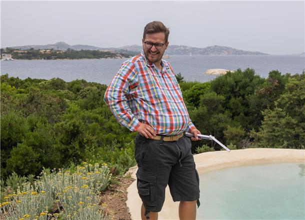 Mann mit Karohemd und kurzer Hose steht an einem Pool. Im Hintergrund sind Bäume, ein größeres Gewässer und Berge zu sehen.