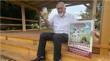 Mann mit Plakat und einem Glas Bier. Gastronom Udo Venema lädt zu einem Brauereifest in sein Erlebnisdorf in Abbehausen ein.