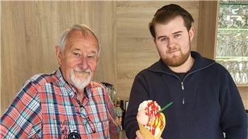 Zwei Männer schauen in die Kamera. Der rechte Mann hält eine große Portion Eis in einer Waffel in die Kamera.