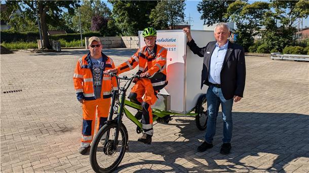 E-Lastenrad hilft bei der Pflege der Spielplätze in Berne Ein Mann in Arbeitskleidung sitzt auf einem großen E-Lastenrad, zu seiner Rechten steht ein Mann ebenfalls in Arbeitskleidung, zu seiner Linken steht ein Mann in Hemd, Sakko und Jeans.