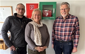Drei Personen stehen lächelnd vor einer weißen Wand mit einem grünen Defibrillatorkasten und einem roten Feuerlöscherkasten.