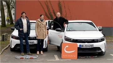 Drei Personen stehen vor zwei weißen Elektroautos von cambio CarSharing. Eine Person lehnt an der offenen Autotür, während die anderen daneben posieren. Ein orangefarbenes cambio-Logo steht vor dem Fahrzeug. Im Hintergrund eine rote Wand mit Efeu.