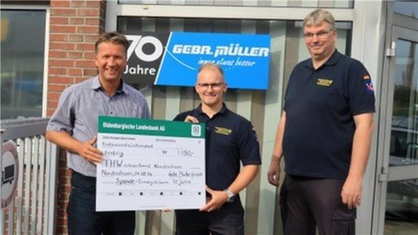 Drei Männer stehen vor einem Fenster, in dem ein Schild mit der Aufschrift "70 Jahre Gebr. Müller" hängt. Zwei der Männer halten einen symbolischen Spendenscheck.