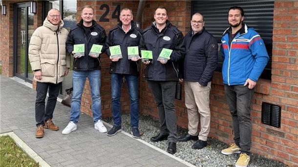 Sechs Männer stehen vor einem modernen Backsteingebäude und lächeln in die Kamera. Drei von ihnen tragen schwarze Jacken mit einem Firmenlogo und halten grüne, gläserne Schilder mit Hausnummern in den Händen. Die Hausnummer „24“ ist an der Wand hinter ihnen sichtbar.