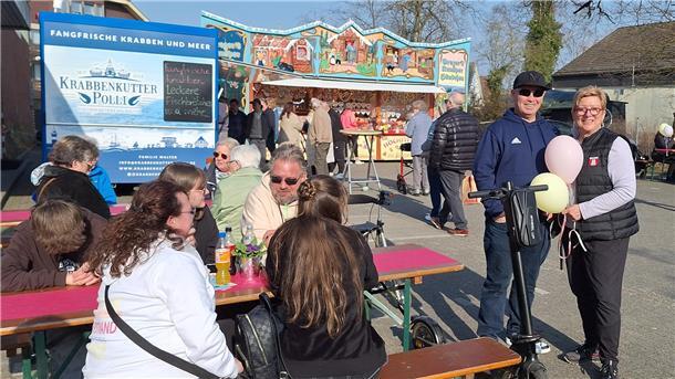 Erste Sonntagsöffnung im neuen Format lockt Hunderte Besucher nach Bad Bederkesa Ein fröhliches Marktgeschehen bei sonnigem Wetter: Menschen sitzen an Biertischen, genießen Speisen und Getränke. Im Hintergrund ein blauer Verkaufswagen „Krabbenkutter Polli“ sowie ein bunt dekorierter Süßwarenstand mit Lebkuchenherzen. Zwei Personen stehen lächelnd mit Luftballons neben einem E-Scooter.