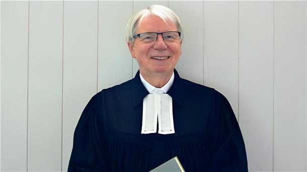 Ein evangelischer Pastor mit Talar und Brille steht vor einer Holztür.